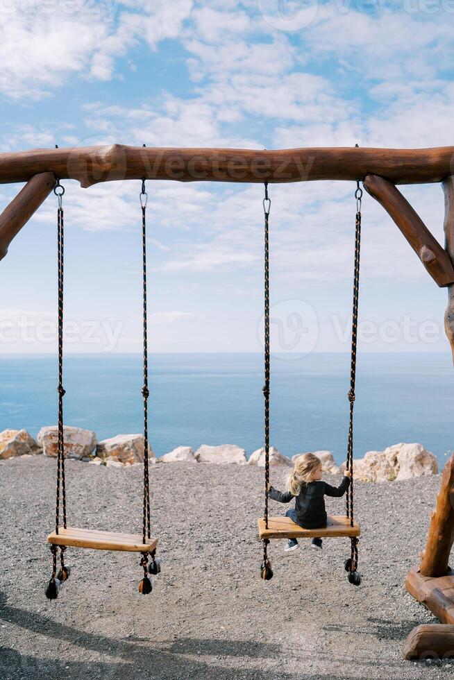 Little girl sits on a large wooden swing on a steep seashore and looks to the side. Back view photo