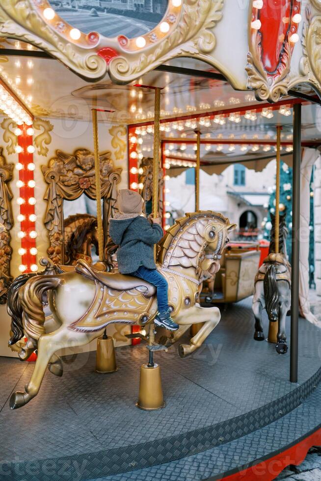 Little girl rides a toy horse on a colorful carousel, holding the handle. Back view photo