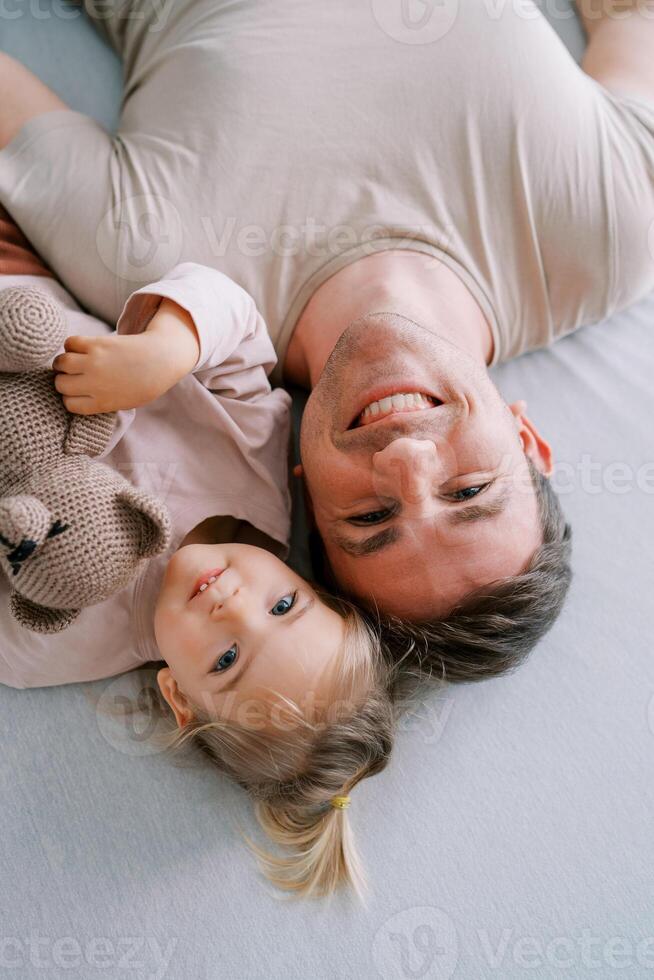 Smiling dad with a little girl and a teddy bear lie on the bed touching their heads. Top view photo