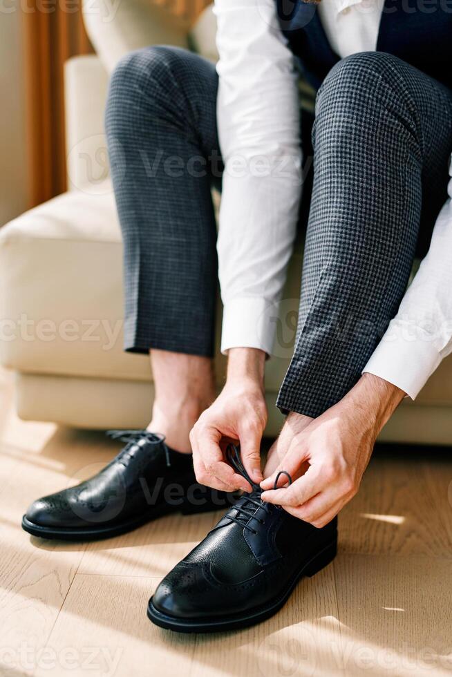 Groom ties his black leather brogues while sitting on the sofa. Cropped. Faceless photo