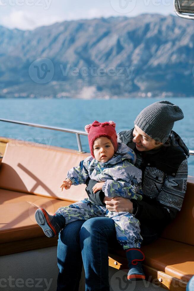 Smiling mother looks at a little girl in her arms sitting in a boat floating on the sea photo