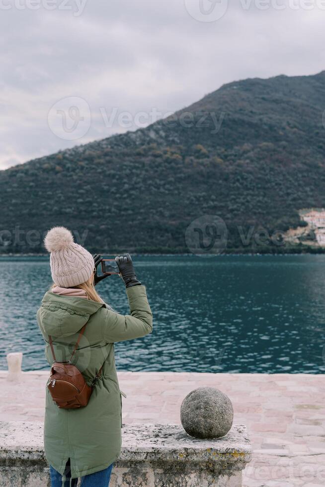 Girl stands by the stone fence on the seashore and takes pictures of the mountains with a smartphone. Back view photo