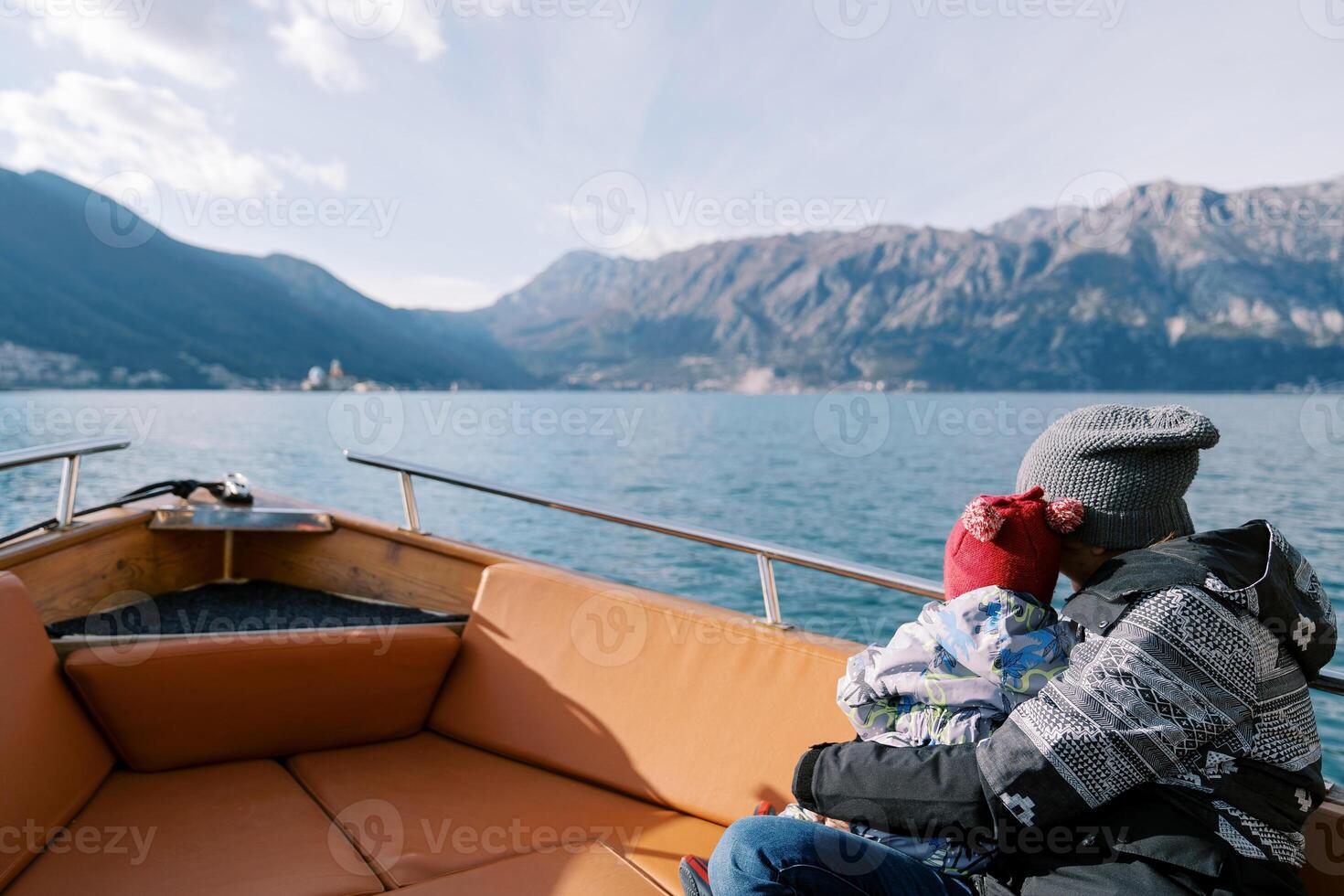 Mom with a small child in her arms floats in a boat along sea and looks at the mountains. Back view photo