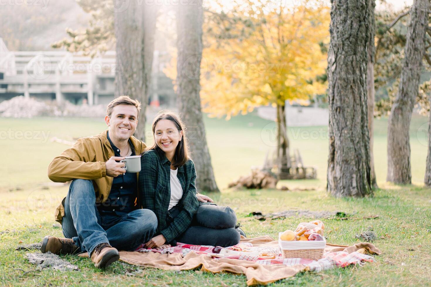 Smiling man with mug hugging woman while sitting on blanket in park near food basket photo