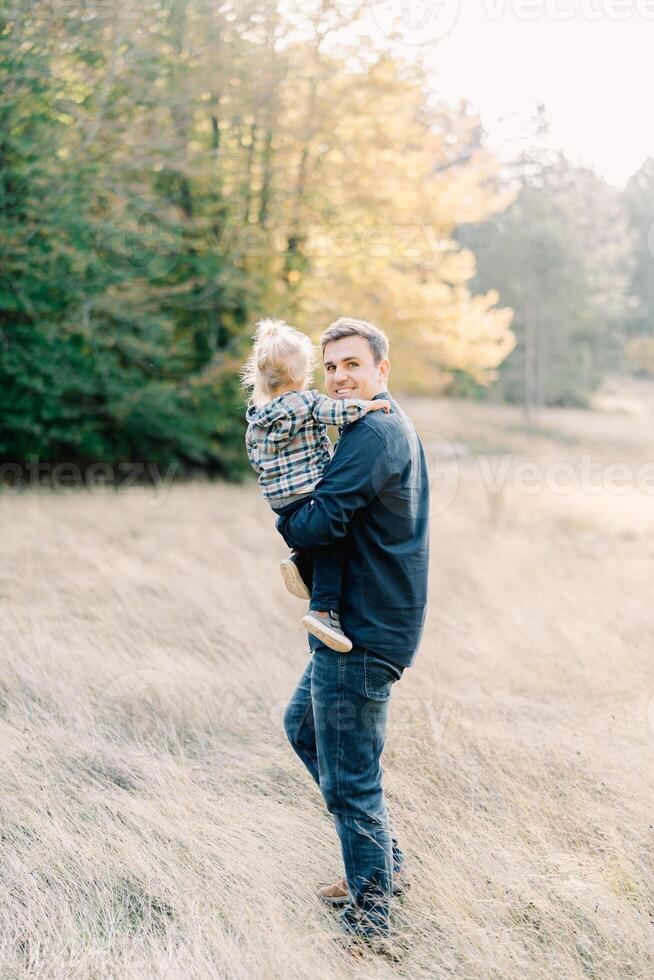 Dad with a little girl in his arms stands in a clearing, looking around. Back view photo