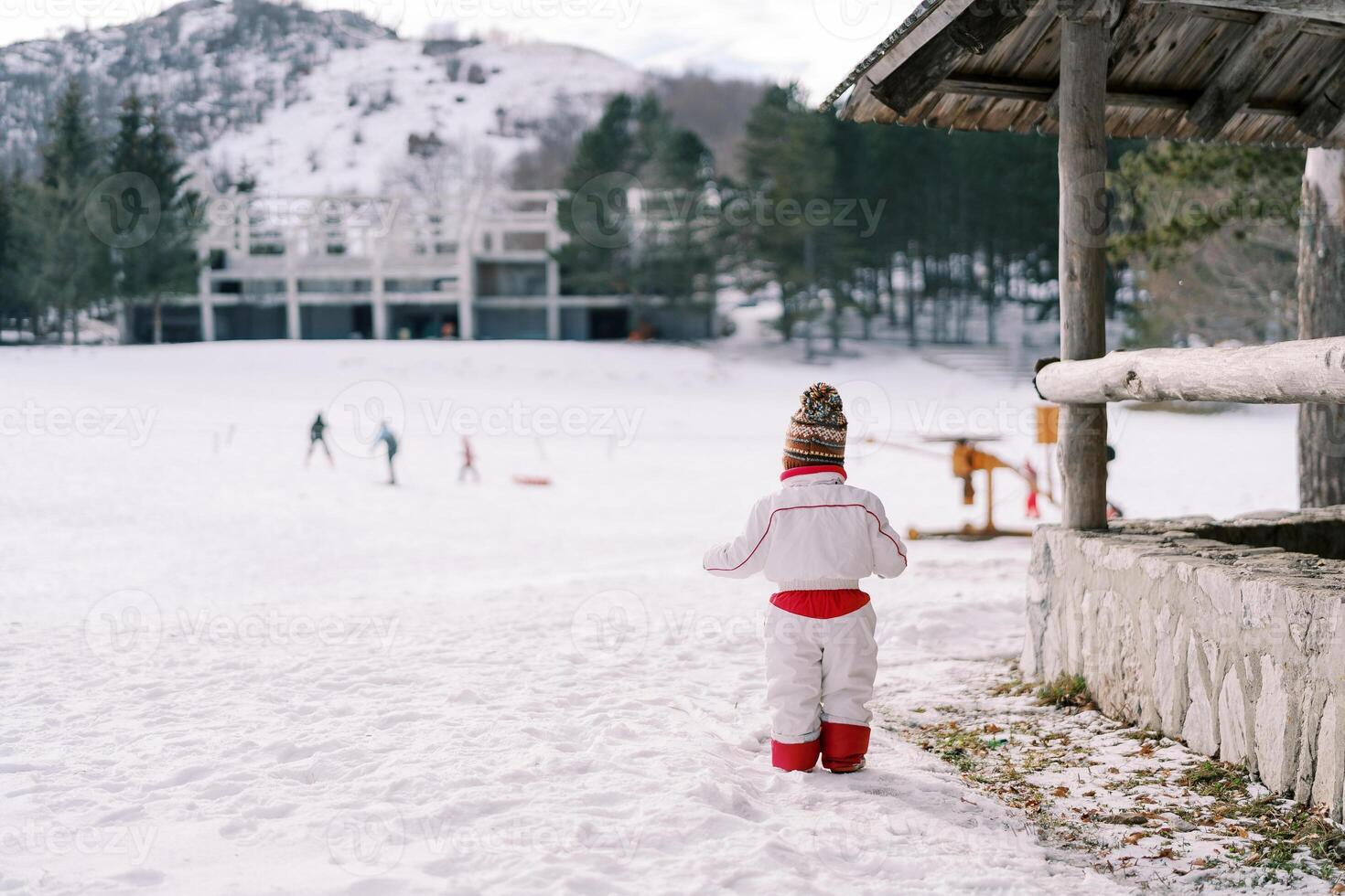 Small child stands near the gazebo on a snowy hill and looks at the skating rink with lugers. Back view photo