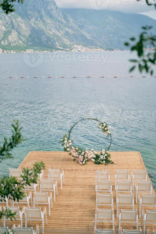 Rows of white chairs stand on the pier in front of a round wedding arch photo