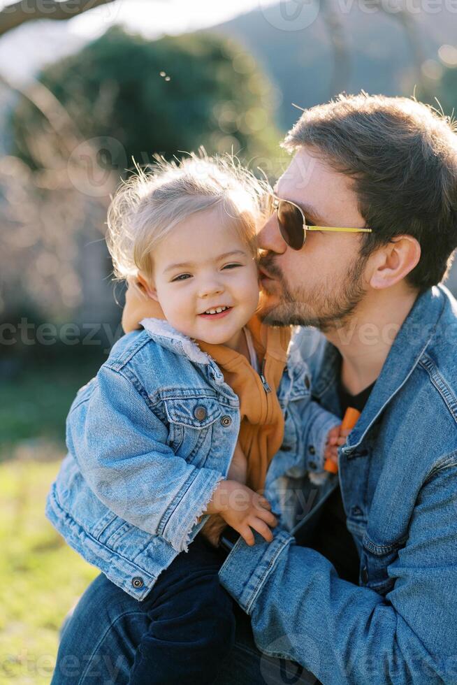 Dad kisses on the cheek a little smiling girl on his knees sitting on the lawn photo