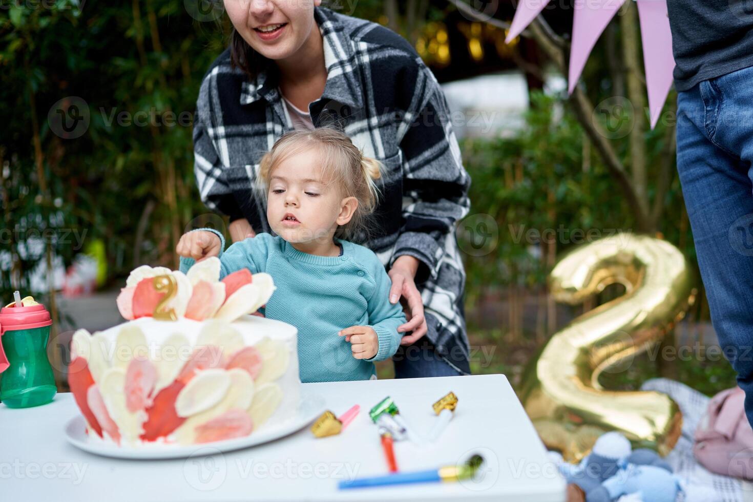 Little girl touches the birthday cake decor on the table with her finger while standing in the garden next to her mother. Cropped photo