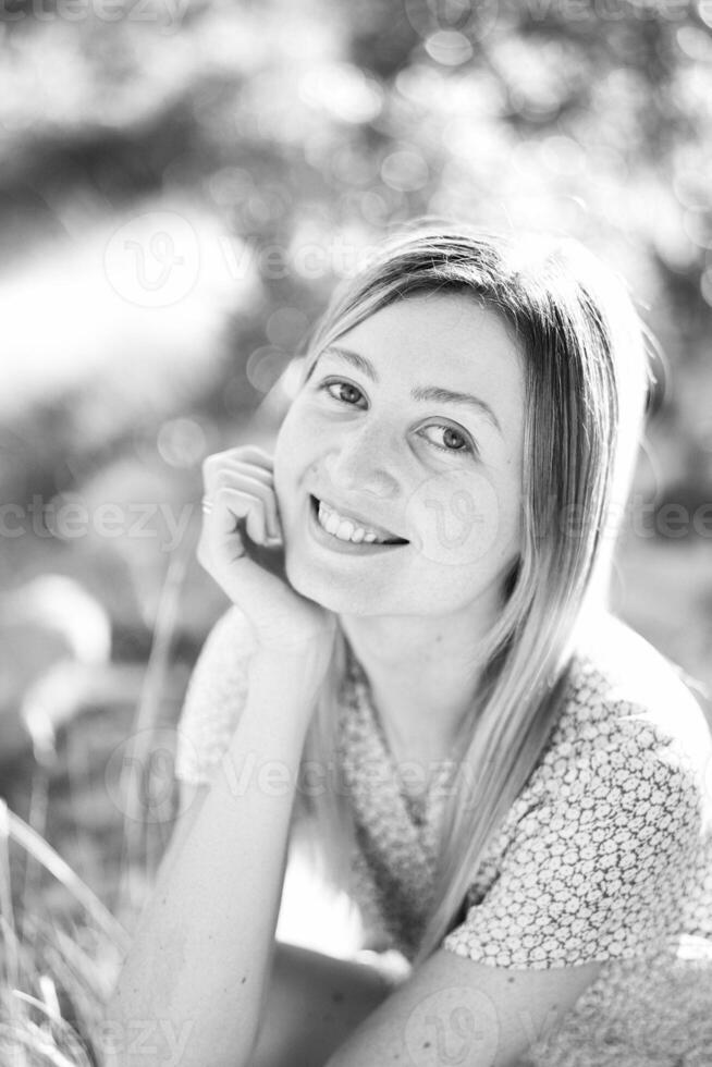 Smiling young woman sits in the grass, resting her head on her hand. Black and white photo