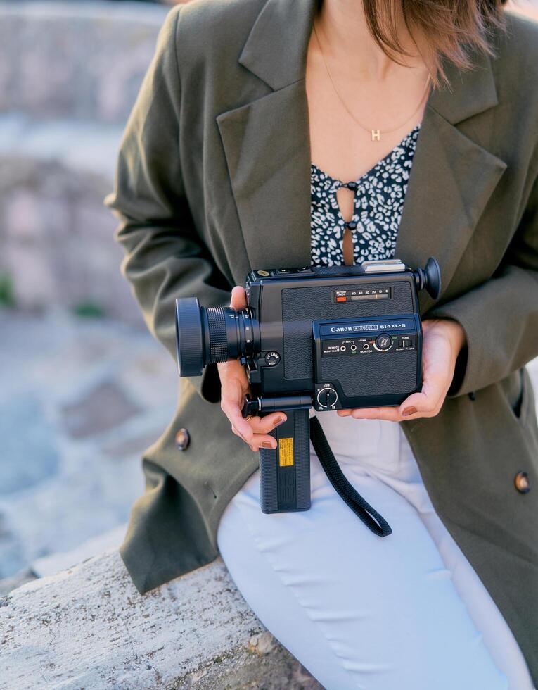 Montenegro, Tivat - July 05, 2024. Girl with a vintage Canon movie camera in her hands sits on a stone fence. Cropped photo