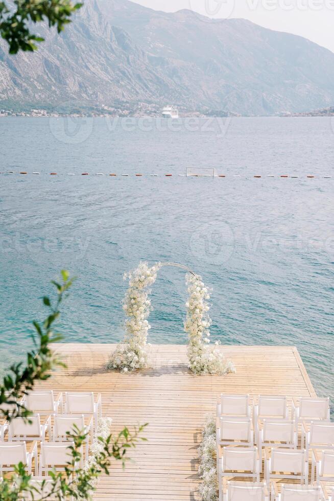 Wedding arch decorated with white flowers stands on a pier in front of rows of white chairs photo