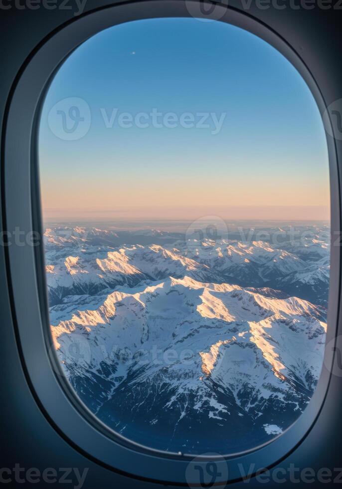 Spectacular snow-covered mountain range unfolds beneath a clear sky, viewed from an airplane window at sunset. photo