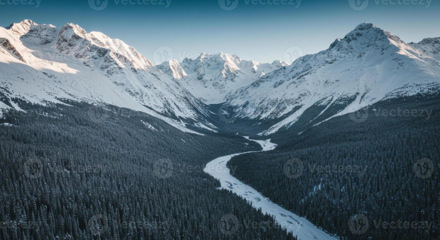 Winding frozen river flows through a vast forest nestled between snow-capped mountain peaks under a clear sky. photo