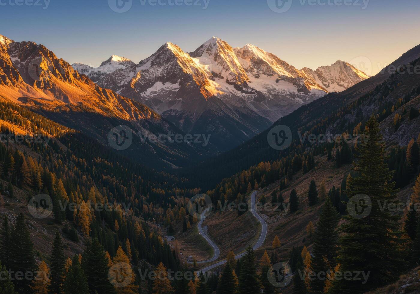 A winding road meanders through a forested valley beneath snow-capped mountains at sunset. photo