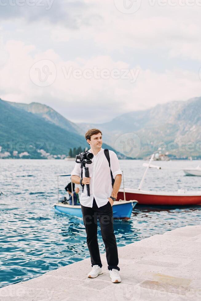 Photographer with a camera on a tripod stands on a pier by the sea and looks into the distance photo