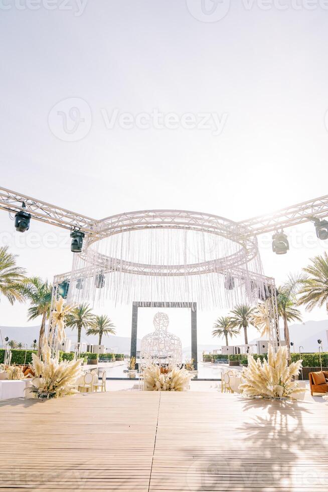 Round stage decorated with bouquets of flowers with spotlights on rectangular stands against the backdrop of palm trees photo