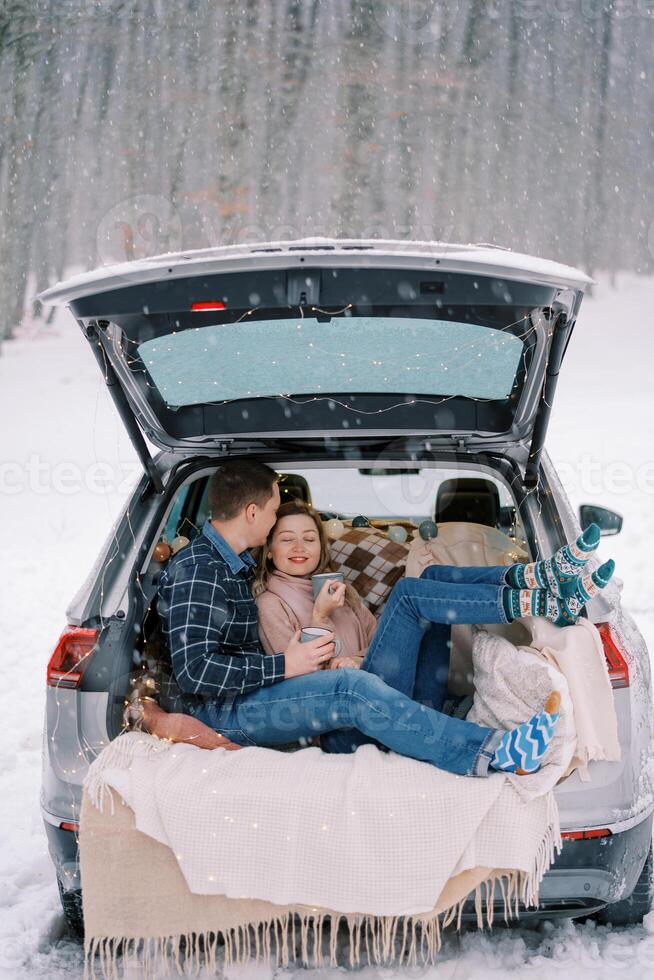 Man kisses a woman on the forehead while sitting in the car trunk with mugs of coffee under snowfall in the forest photo