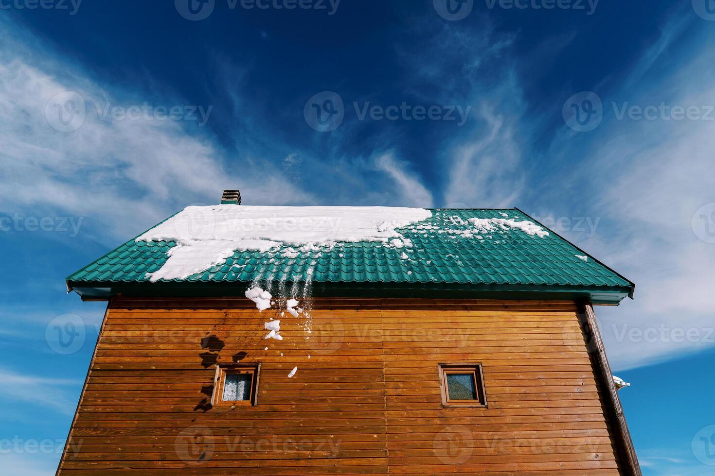 Snow falls from the green tiled roof of a wooden cottage against the blue sky photo