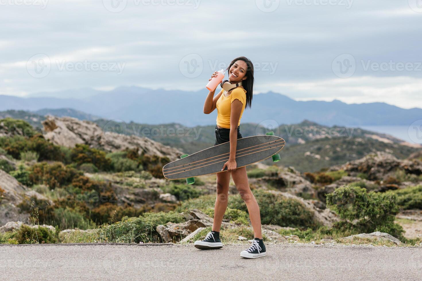 Smiling skater with longboard walking on pavement against mount photo