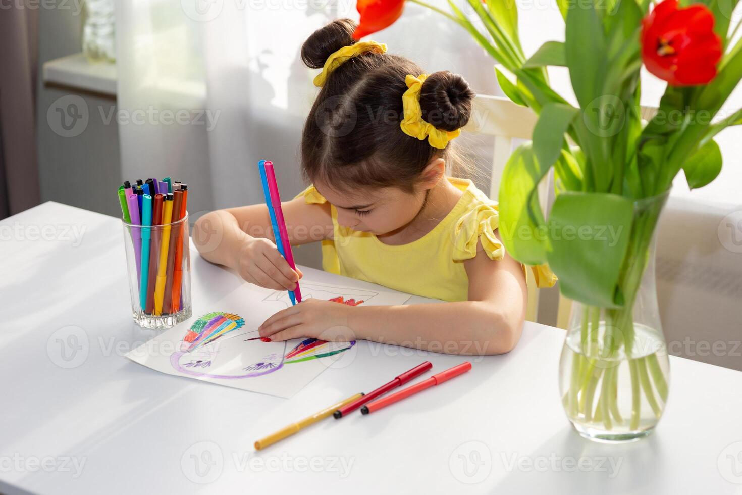 Cute little brunette girl in yellow dress , drawing with colored markers on the white table photo
