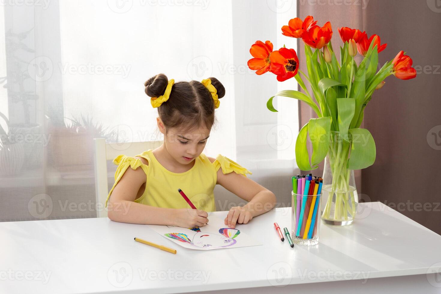 Cute little brunette girl in yellow dress drawing with colored markers on the white table in home photo