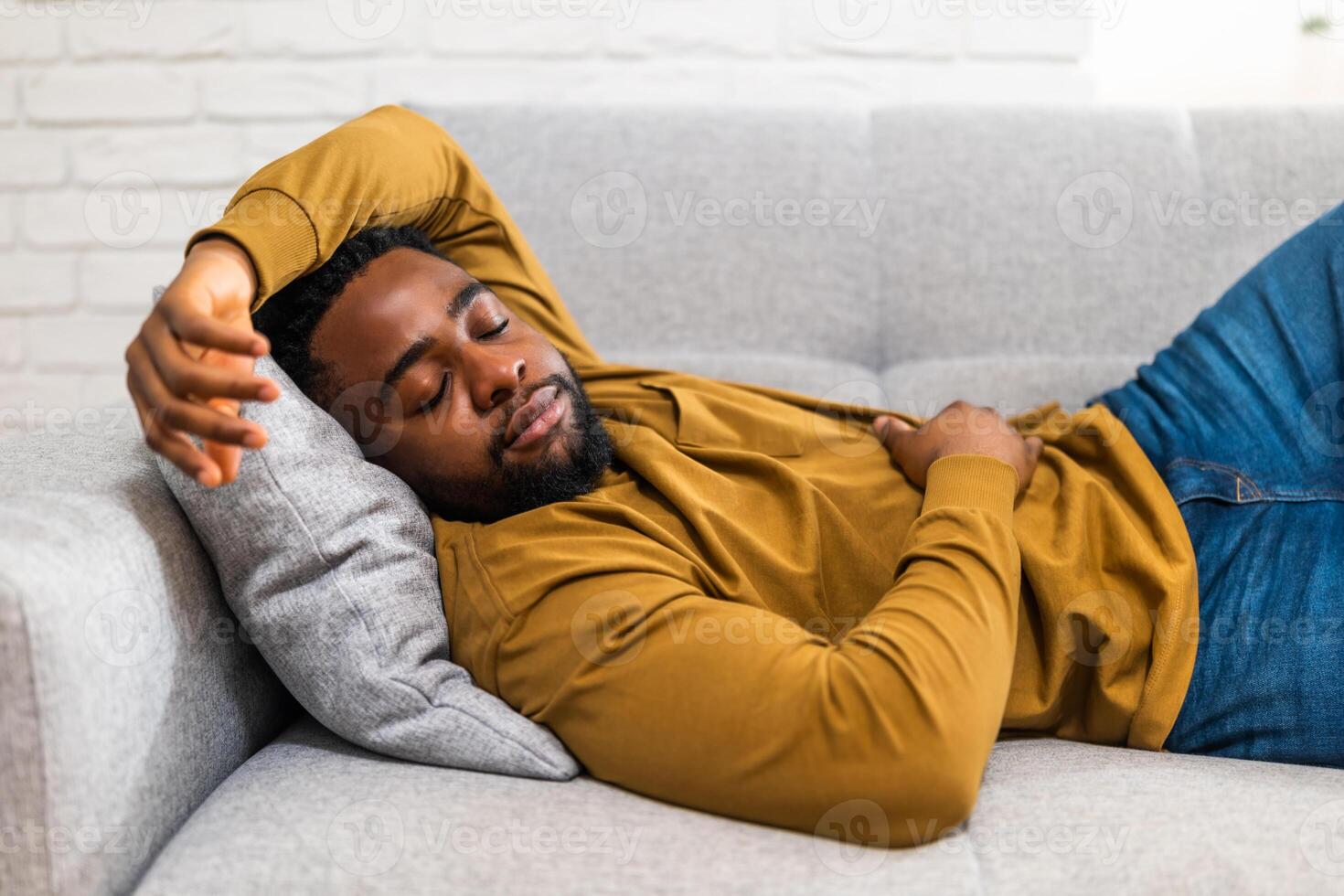 Serene African American man in casual clothing enjoying in an afternoon nap while lying comfortably on sofa in his modern living room. photo