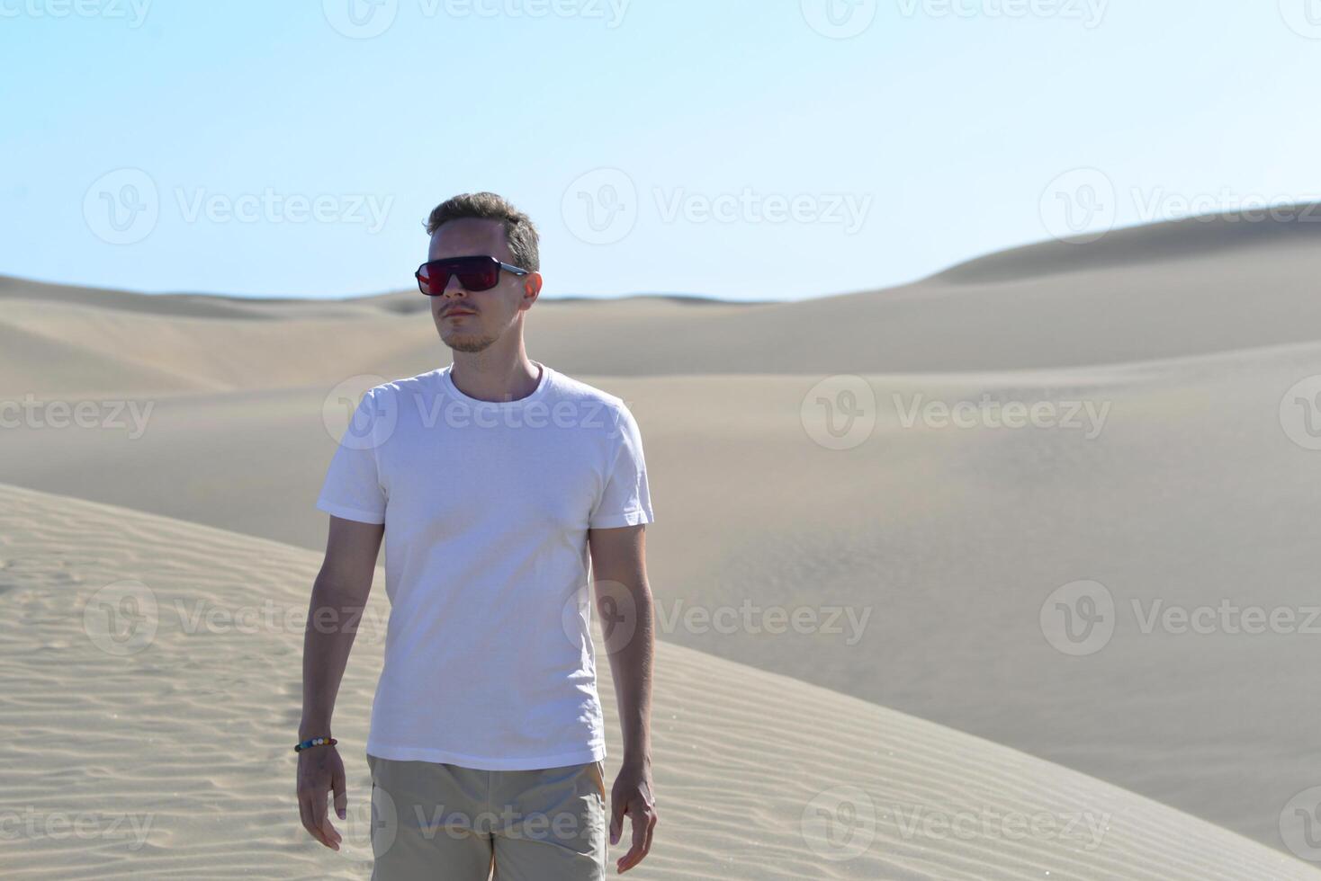 A stylish model portrait of a young man walking along a sand dune through barkhan in Maspalomas, Gran Canaria on a hot sunny day. Desert travel touristic concept photo