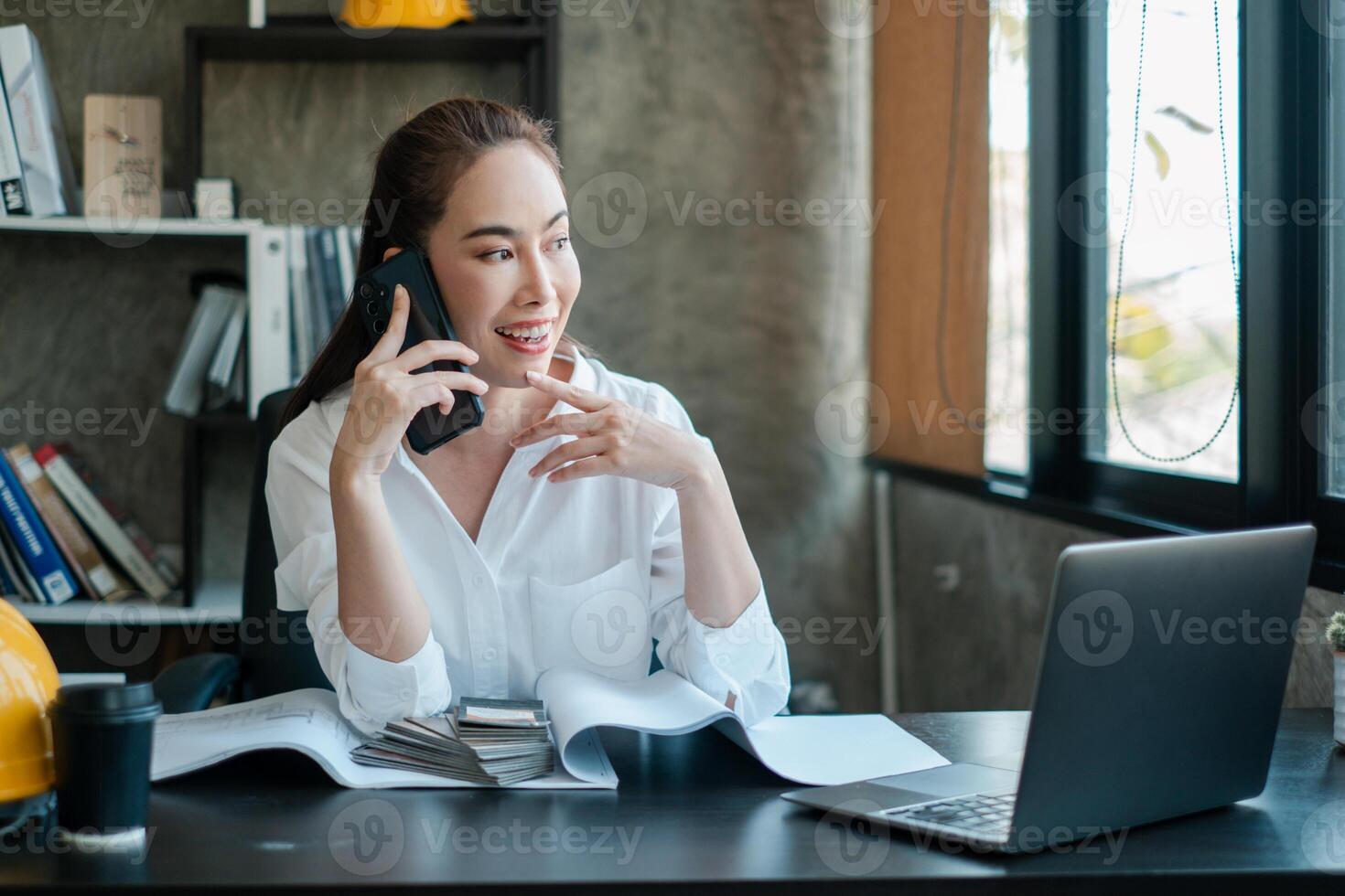 Businesswoman multitasking in a modern office, talking on the phone, using a laptop, and reviewing documents. Bright and productive workspace. photo