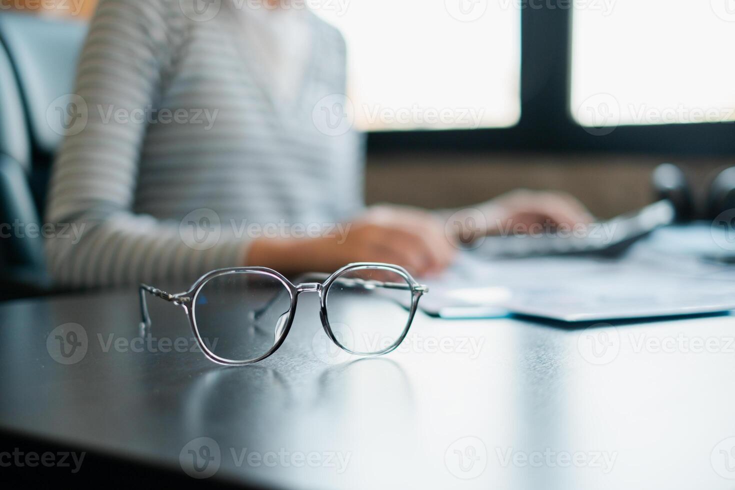 Modern eyeglasses on a desk with a blurred person working on a computer, symbolizing focus and productivity. photo