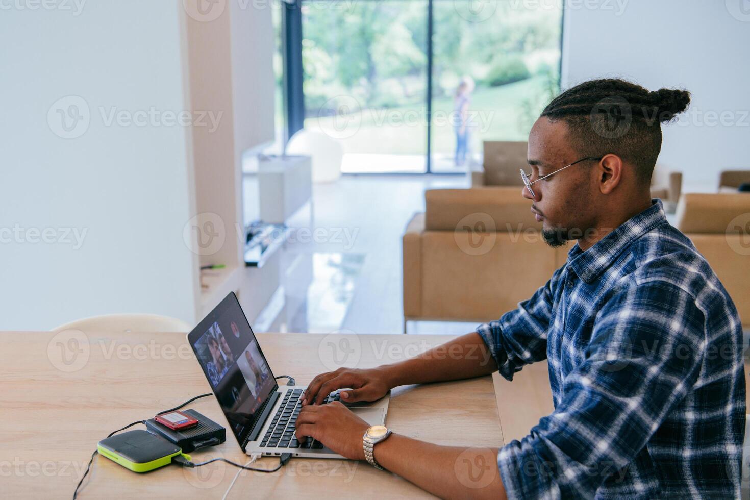 African American influencer in glasses sitting at a table in a modern living room, using a laptop for business chat, conversation with friends and entertainment. photo