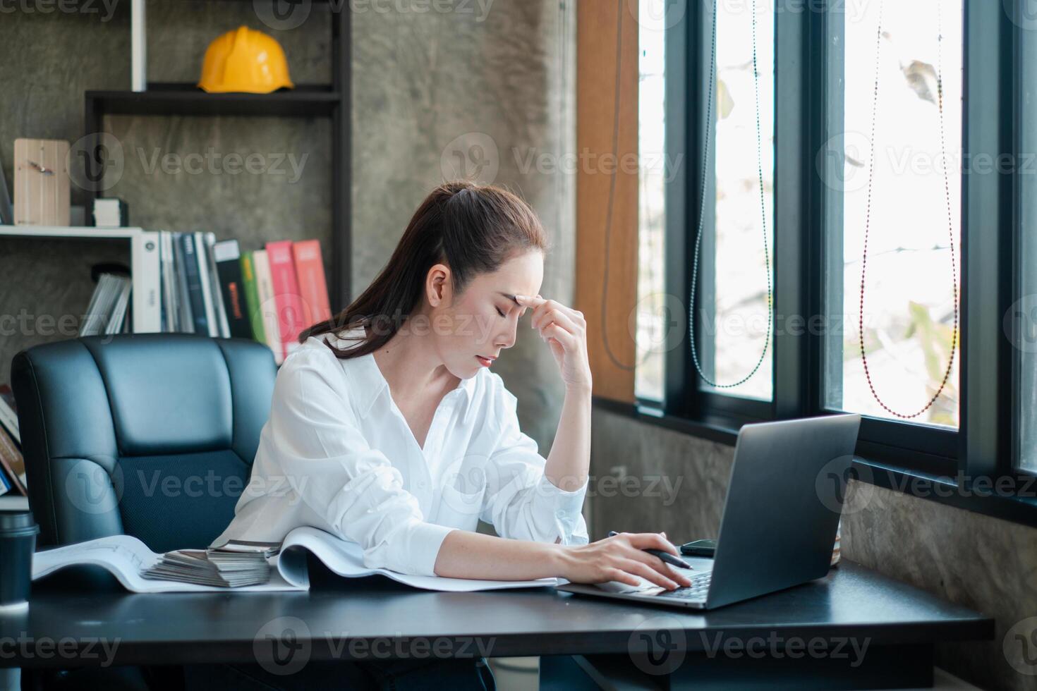 A businesswoman in a modern office, stressed while working on a laptop with architectural plans and a safety helmet nearby. photo