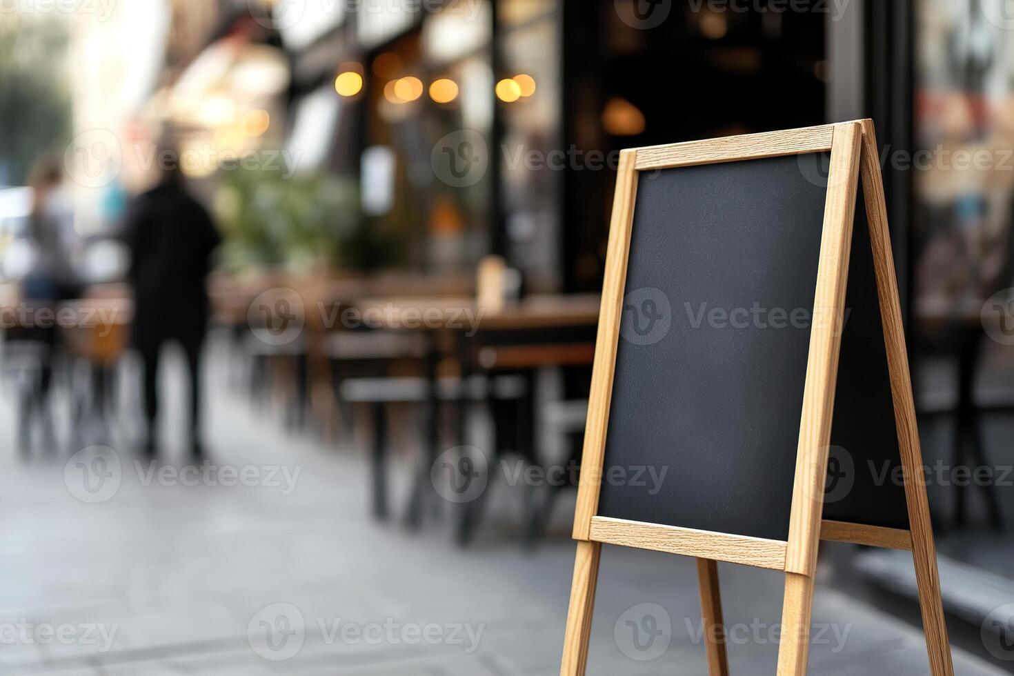 A blank A-frame sandwich board stands prominently on a bustling sidewalk outside a trendy cafe. The softly blurred background reveals patrons at wooden tables and a glass storefront photo
