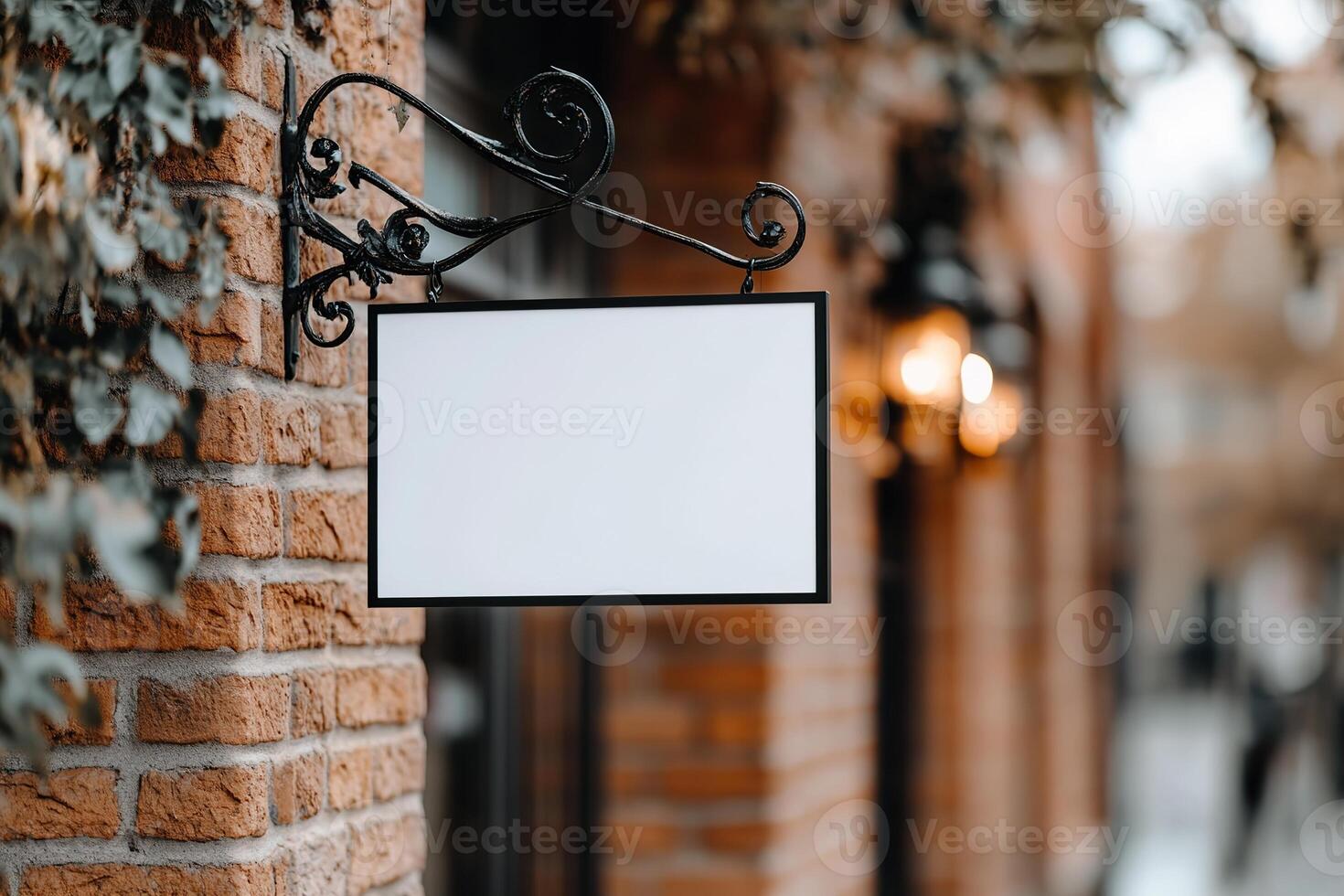A blank rectangular signboard hangs from an ornate wrought iron bracket on a vintage brick wall, evoking a nostalgic atmosphere in a charming European street with warm lighting photo