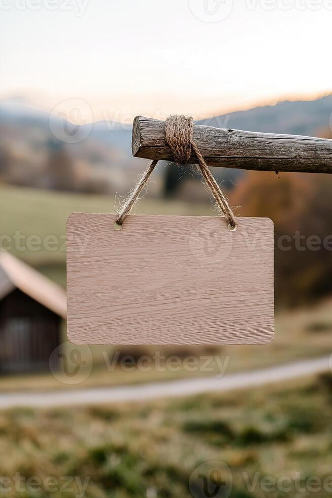 A blank wooden signboard hangs from a wooden post in a serene rural setting. The softly blurred background features rolling hills, a barn, and a dirt road, providing a tranquil atmosphere photo