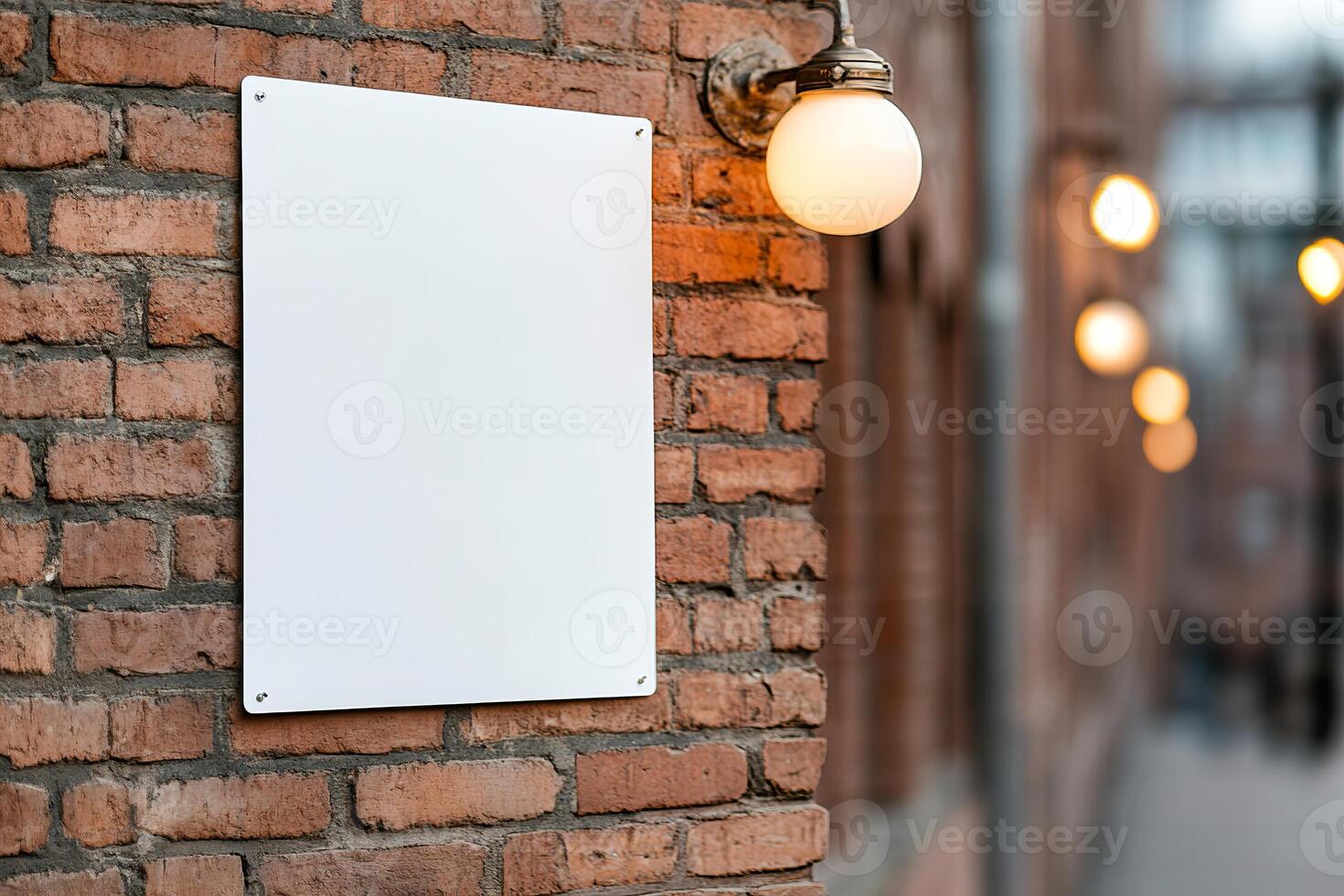 An empty billboard is mounted on a weathered red brick wall in a historic downtown area. Softly glowing street lamps illuminate the surroundings during the warm golden hour photo