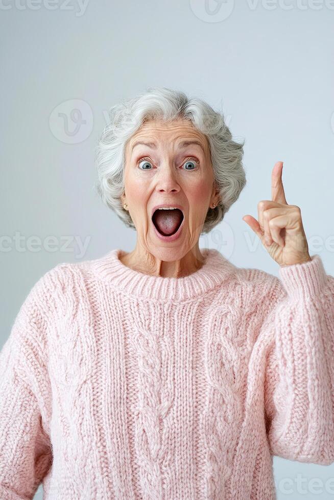 A lively senior woman wearing a casual sweater shows her surprise with an open mouth and excited gesture. The clean white background enhances her energetic expression and enthusiasm photo