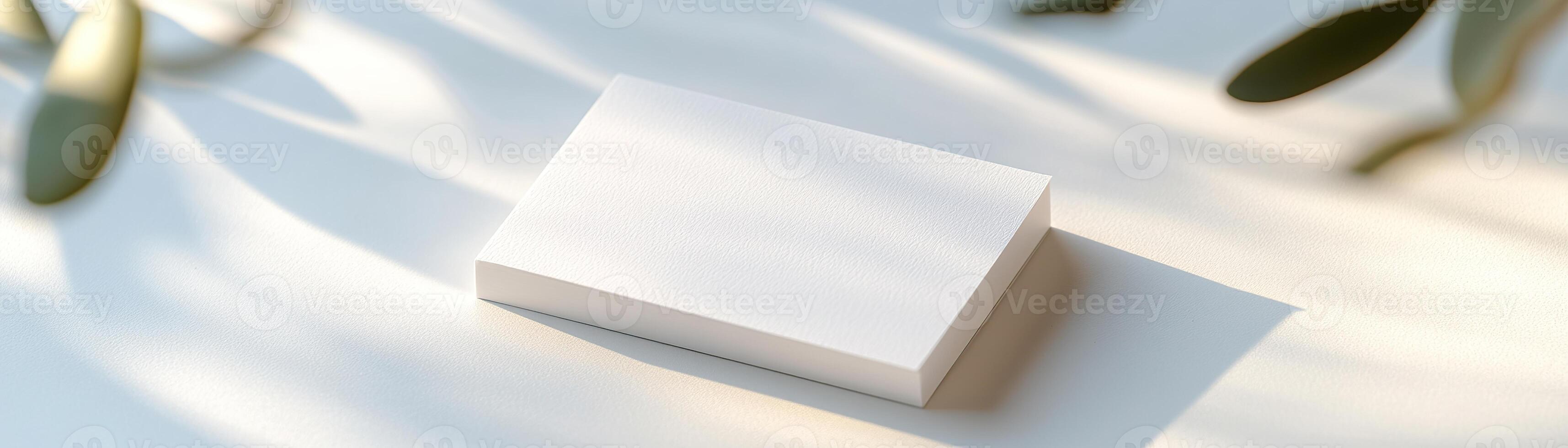 A blank business card rests on a smooth white surface, softly lit by natural light that creates gentle shadows. The background is blurred, highlighting the card's clean design and professional appeal photo