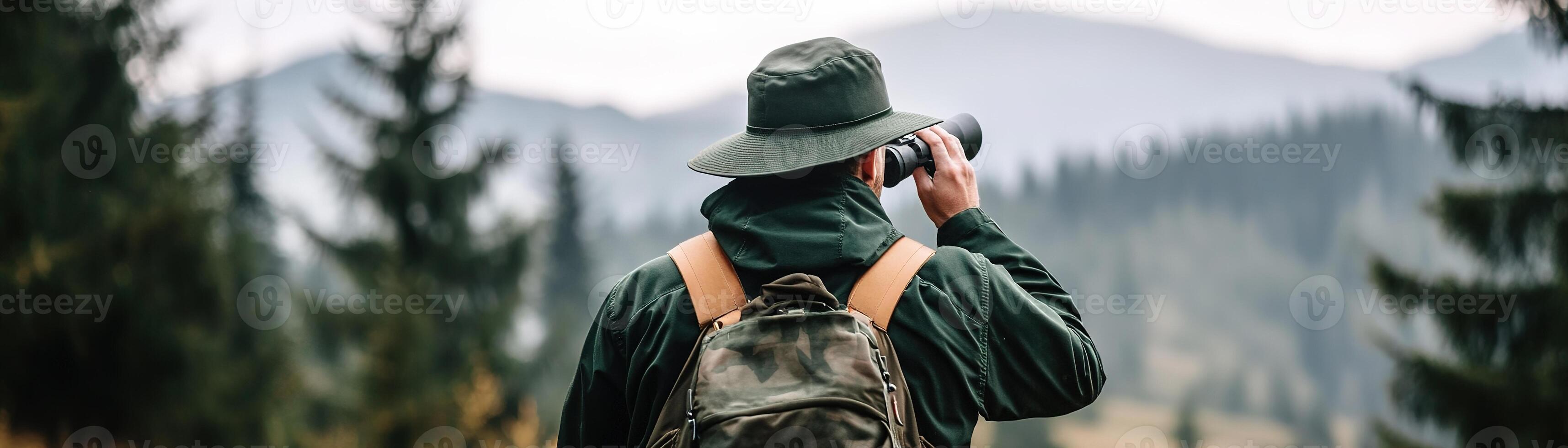 A park ranger in a green uniform stands on a beautiful hiking trail, holding binoculars and observing the stunning mountain scenery and tall pine trees around him photo