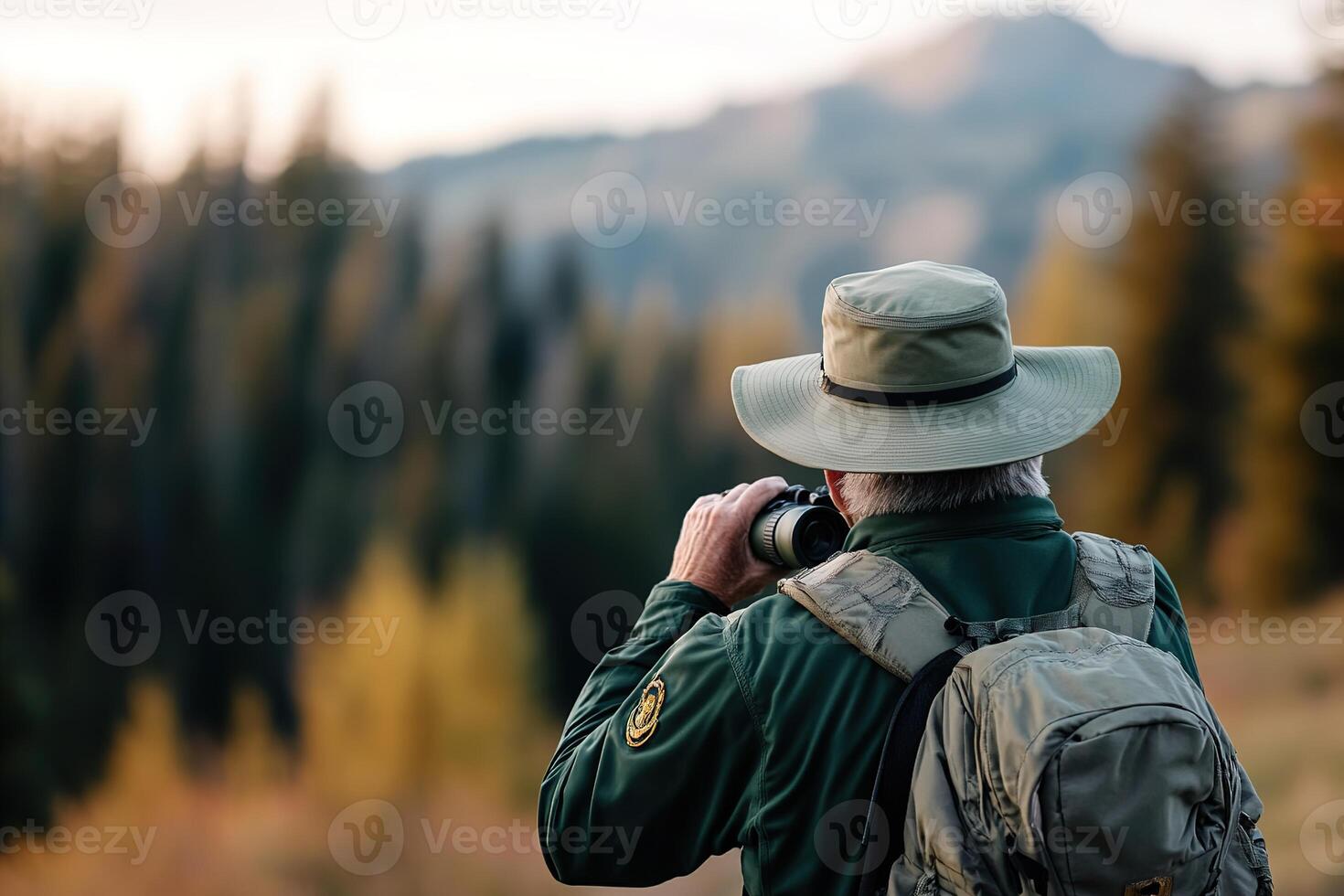 A park ranger in a green uniform stands on a beautiful hiking trail, using binoculars to observe wildlife amidst tall pine trees and a stunning mountain backdrop photo