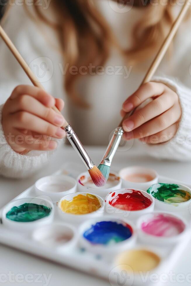 Hands of a child joyfully dip brushes into bright watercolor paints, exploring creativity and color at a table. Soft focus enhances the artistic atmosphere, inspiring early education photo