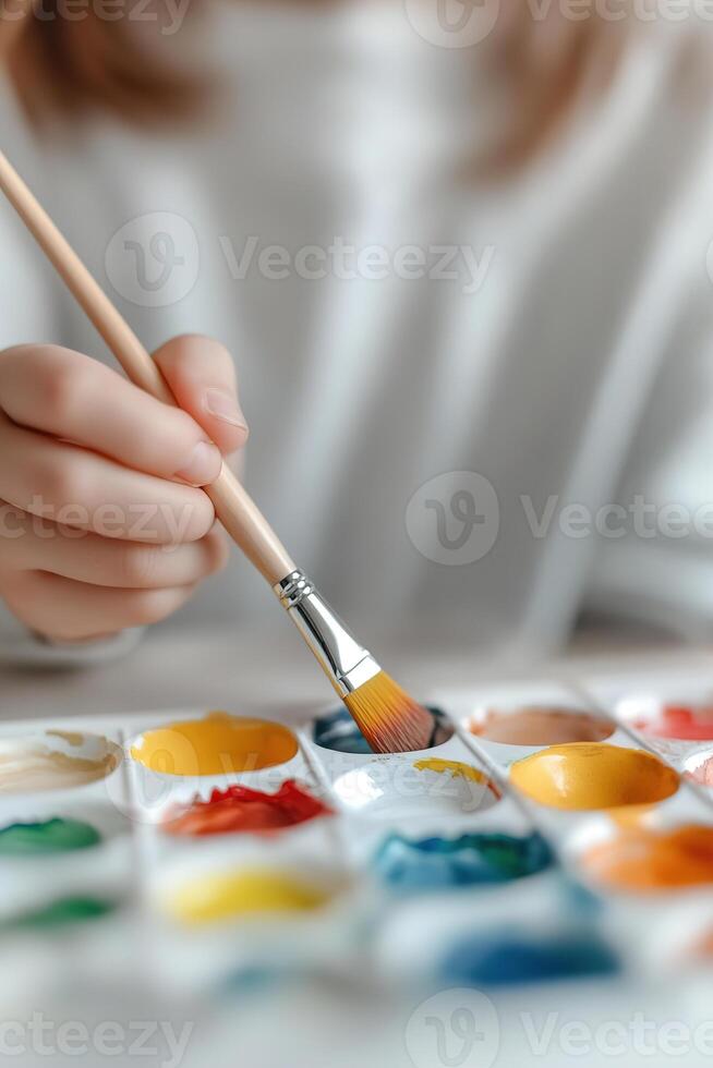A close-up view captures small hands dipping brushes into bright watercolor paints, engaged in an artistic activity on a table in a softly lit environment, perfect for early learning photo