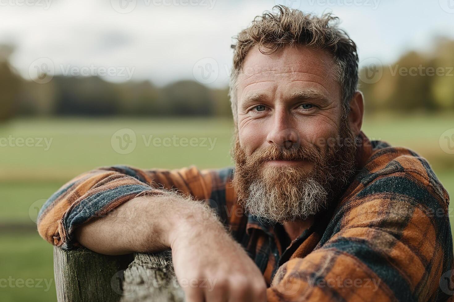 A rugged farmer with a thick beard and flannel shirt leans thoughtfully on a wooden fence, surrounded by softly blurred green fields and trees, embodying rural tranquility and sustainability photo