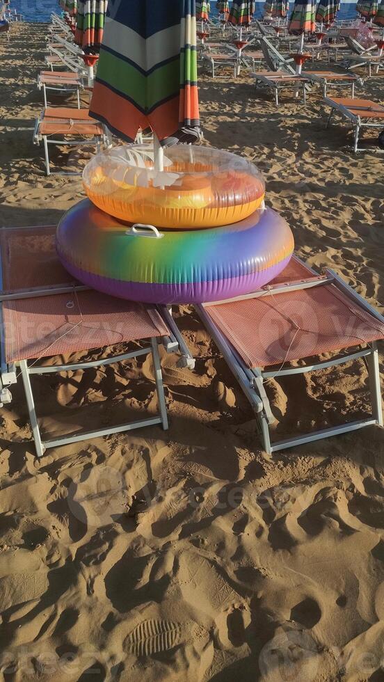 Colorful inflatable rings resting on beach chairs beneath umbrella, sandy shoreline of bibione, italy, evoking summer leisure photo