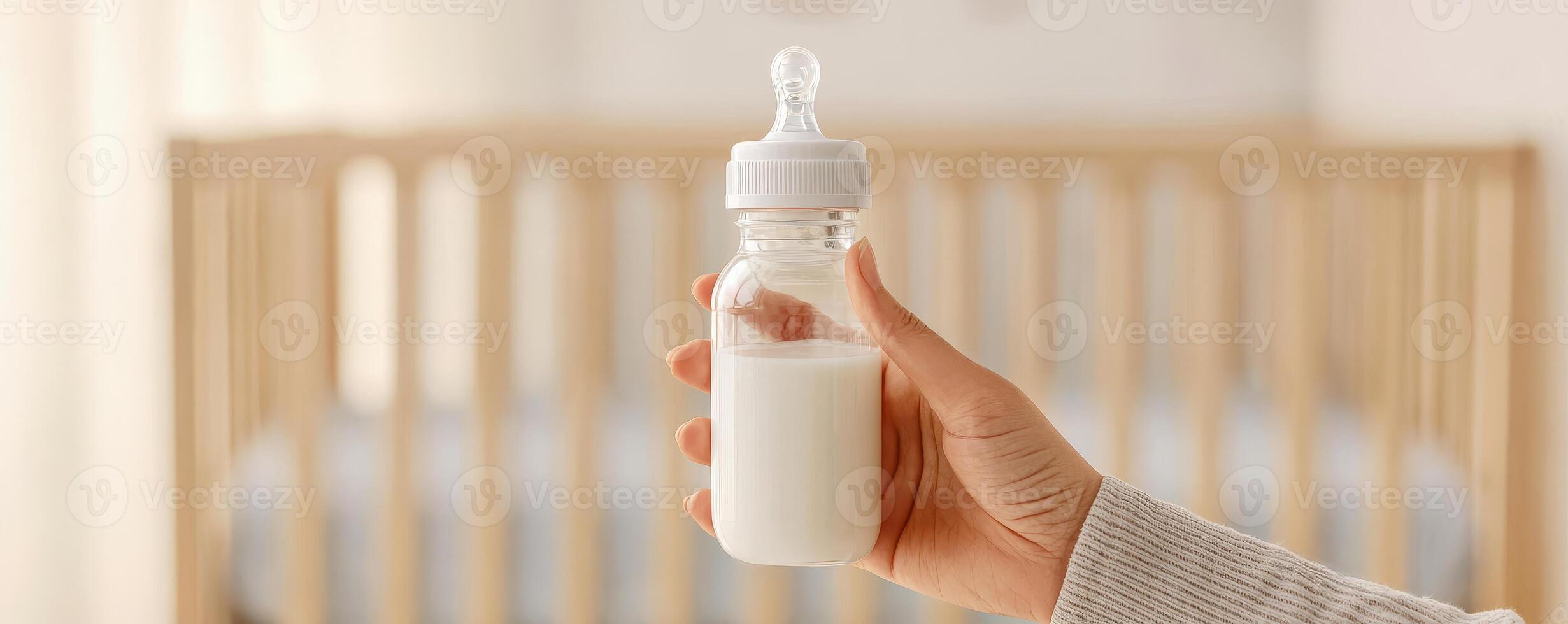 Parenting resources for family concept. Hand holds a baby bottle filled with milk, set against a soft-focus background of a nursery with a crib. photo