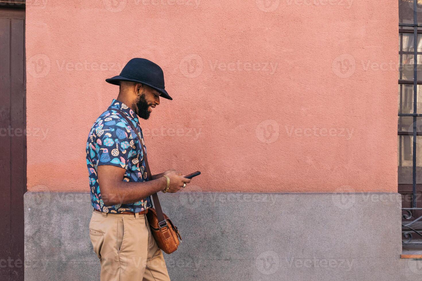 A man in a hat and shirt standing next to a wall looking at his phone photo