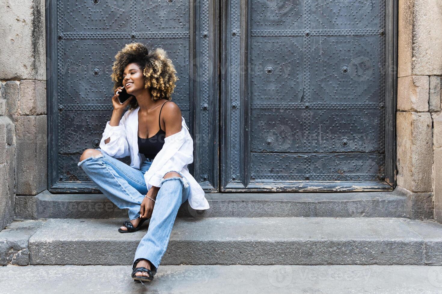 Delighted black woman speaking on smartphone while sitting near building photo
