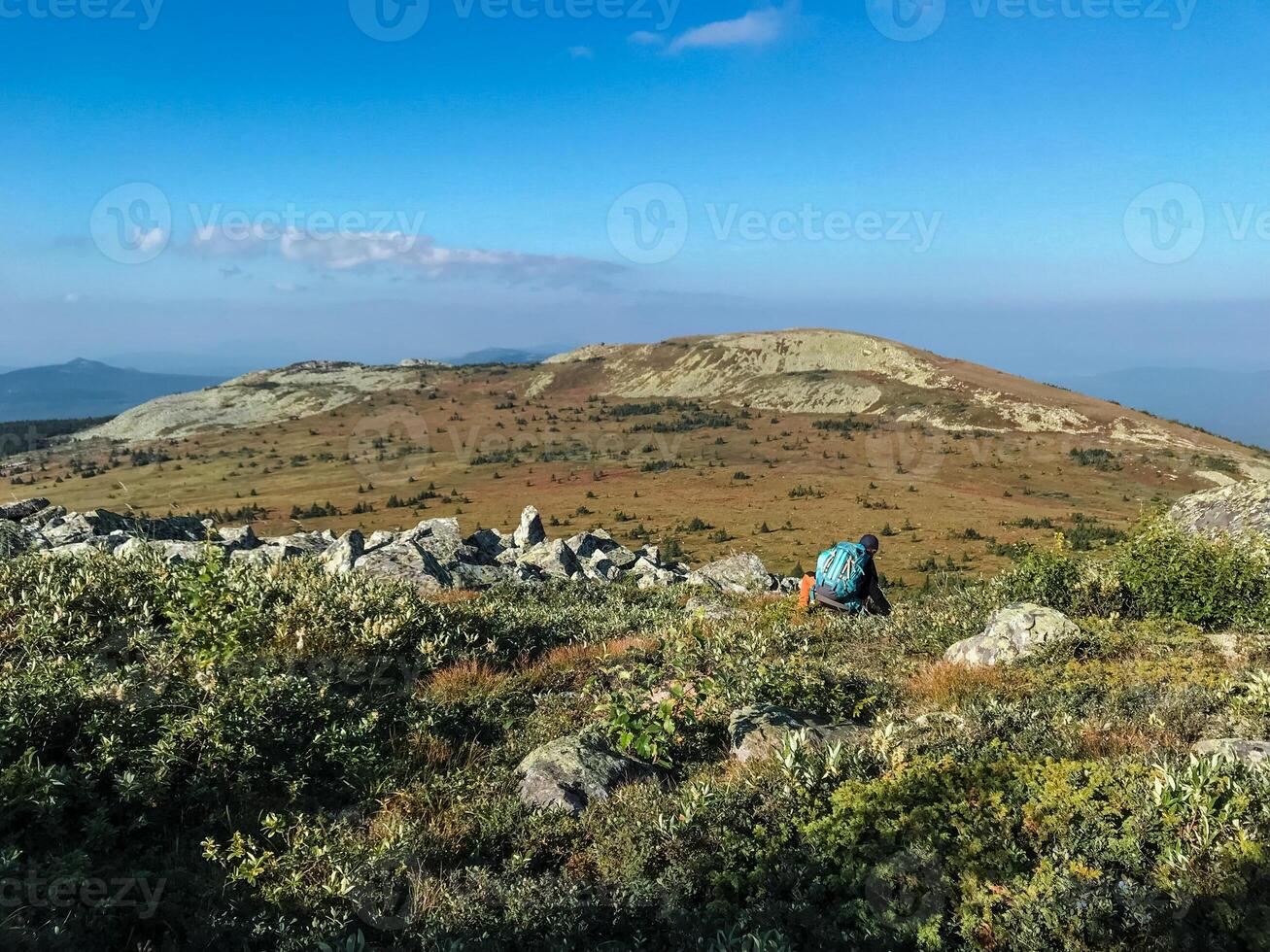A solitary individual sits calmly on a scenic hilltop with a sturdy backpack, enjoying the breathtaking beauty and tranquility of the surrounding highlands and shrubland photo