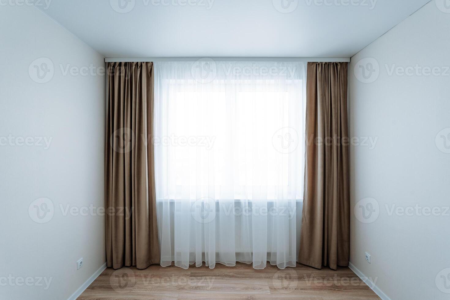 The room is completely empty, featuring a large window dressed with brown curtains that gently sway in the soft breeze, providing a sense of openness and light to the space photo