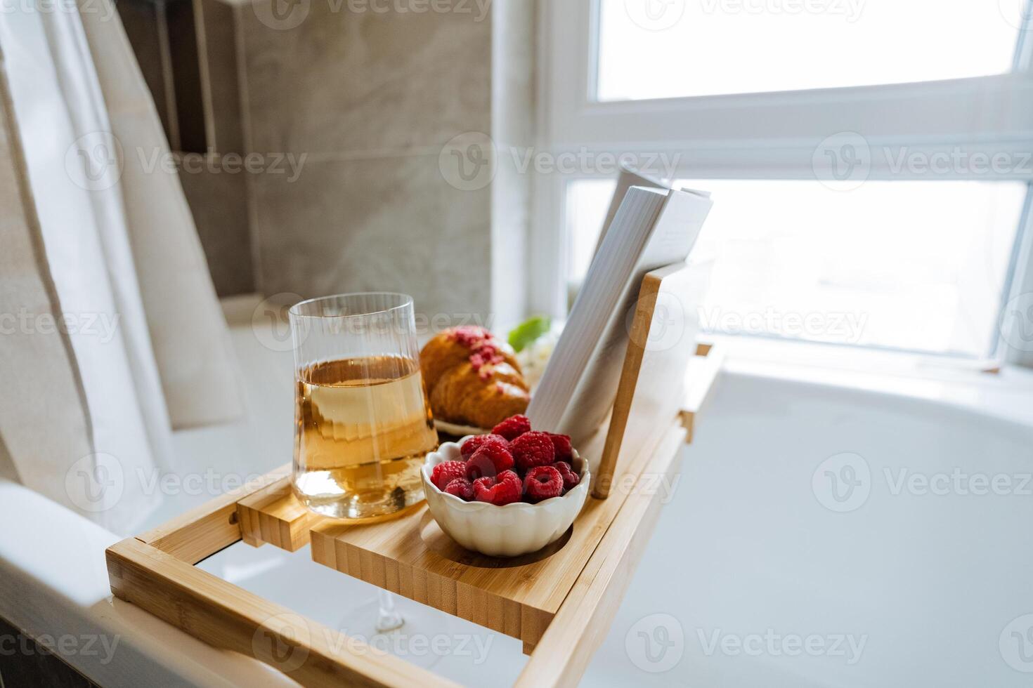 A beautifully crafted wooden bathtub tray is adorned with fresh, plump raspberries, a flaky croissant, a glass of fine wine, and an engaging book to read during a relaxing bath photo